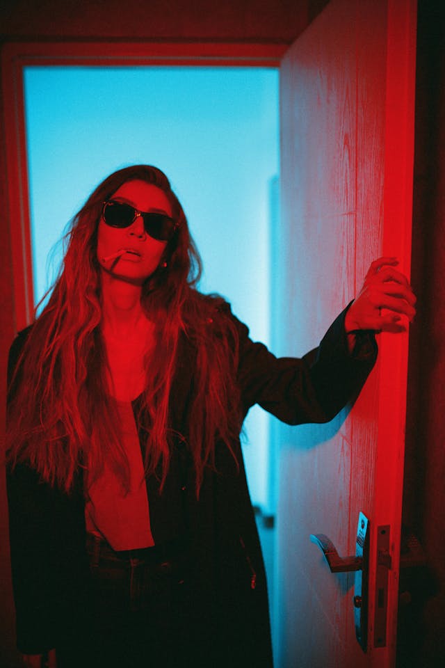 Woman in doorway lit by dramatic red and blue lighting, wearing sunglasses and a dark coat, cigarette in mouth—evoking a moody, cinematic neo-noir atmosphere.